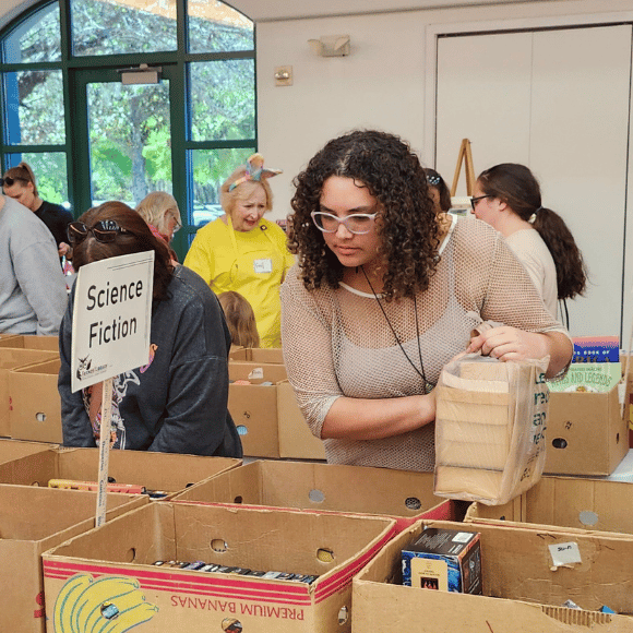 Browsing the booksale Women holding plastic bag filled with books glances into a box of science fiction books